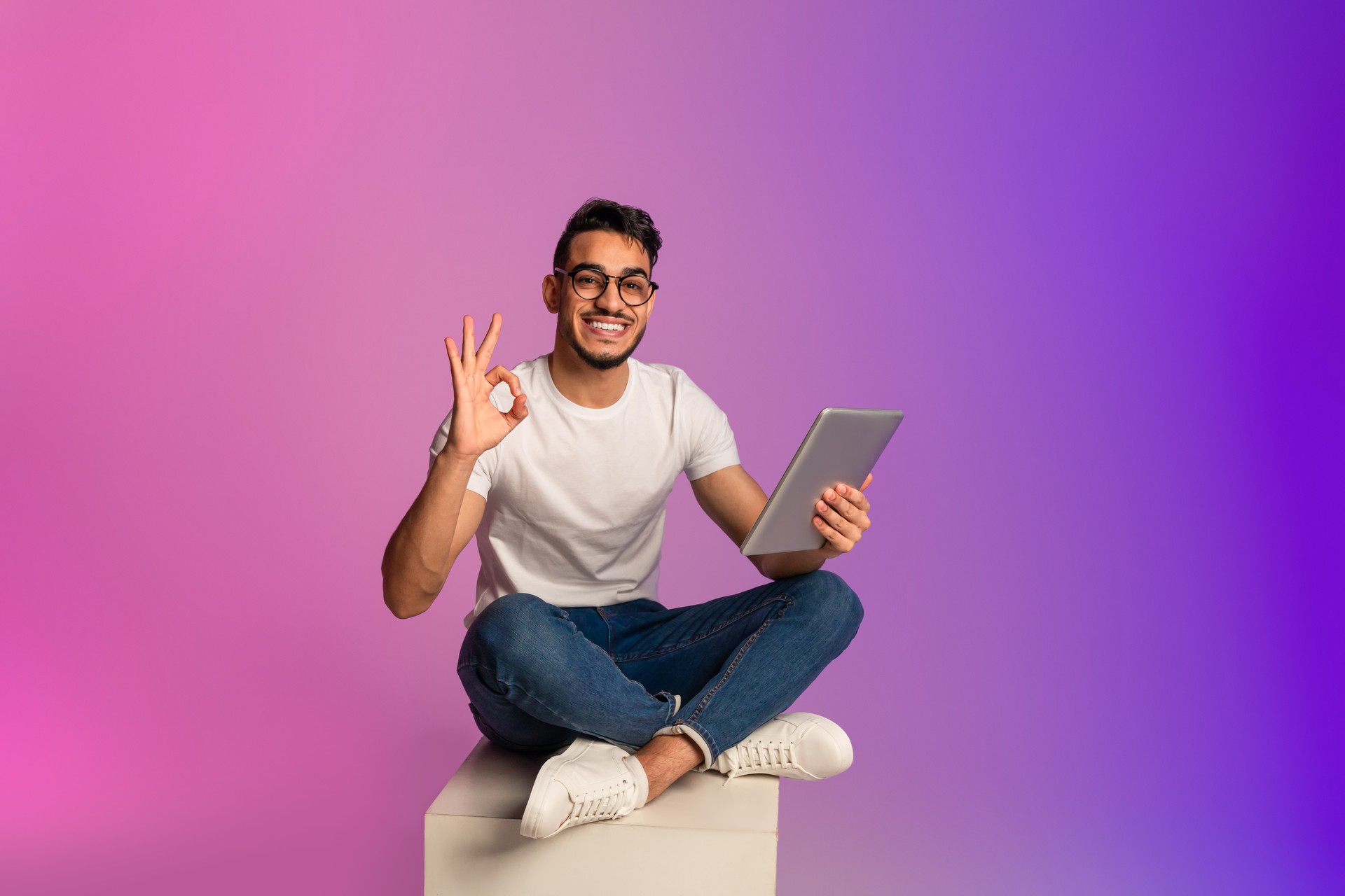 Full length of young Arab guy using tablet pc, showing okay gesture, recommending new app in neon light